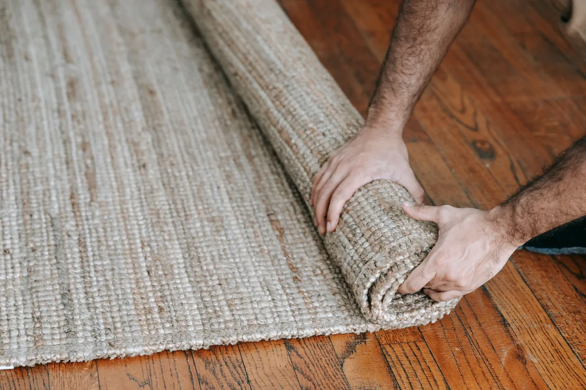 Two hands rolling up a chunky woven natural-fiber rug on a wooden floor