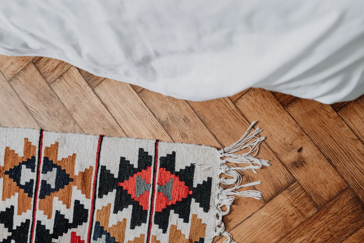 The corner of a patterned flatweave rug with a fringed edge on a herringbone wood floor beside a bed with white linen