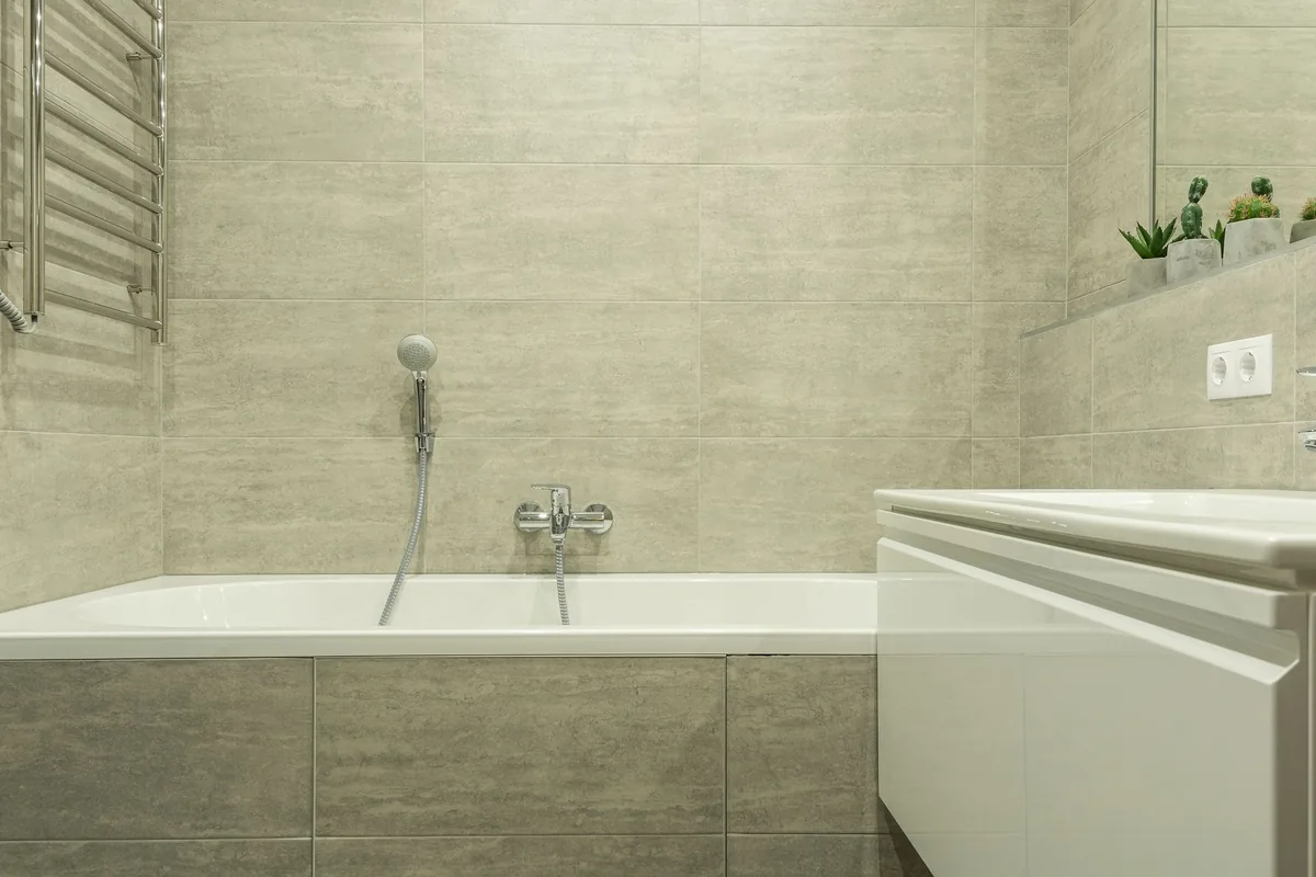 A white bathtub with a handheld shower and wall tap against gray stone-look wall tiles, beside a white wall-hung vanity