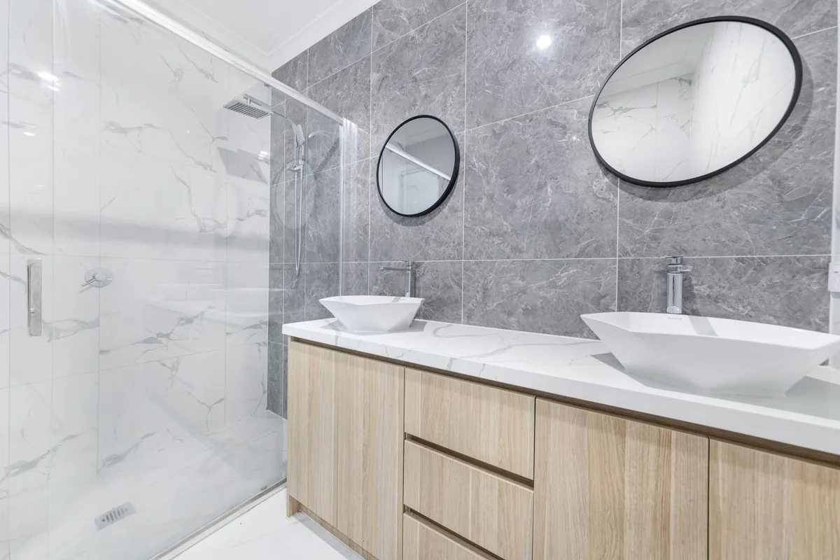 A glass shower enclosure beside a wood-front double vanity with two vessel sinks and oval mirrors on gray marble-look wall tiles