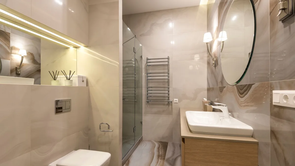 Bathroom with large marble-look wall tiles, a glass shower, a wall-hung toilet, a round mirror and a wood vanity with a white basin