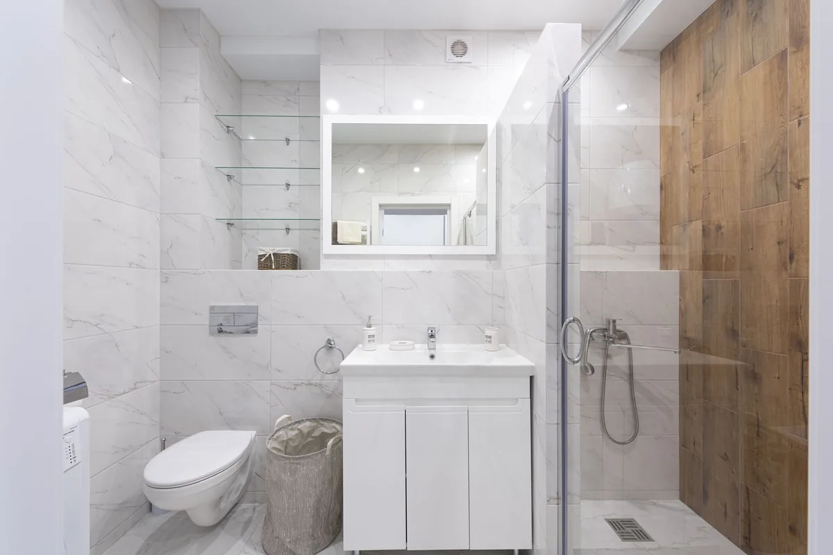 A small marble-look bathroom with glass shelves above a wall-hung toilet, a mirror, a small vanity and a glass shower