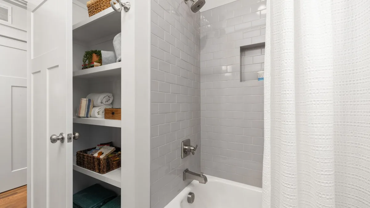 A white bathroom with open built-in shelves of towels and baskets beside a tub and a tiled shower niche