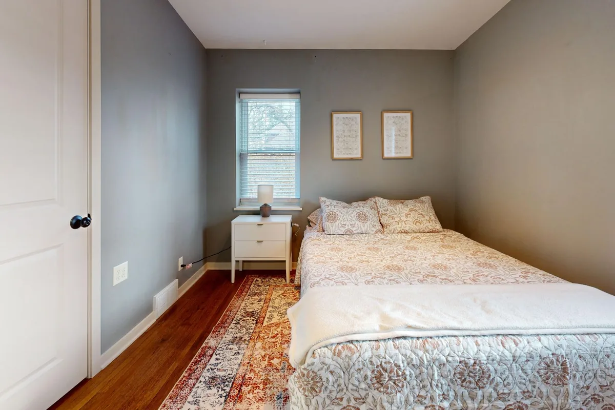 A small bedroom with a bed set against one wall, a white bedside table under the window and a rug beside the bed