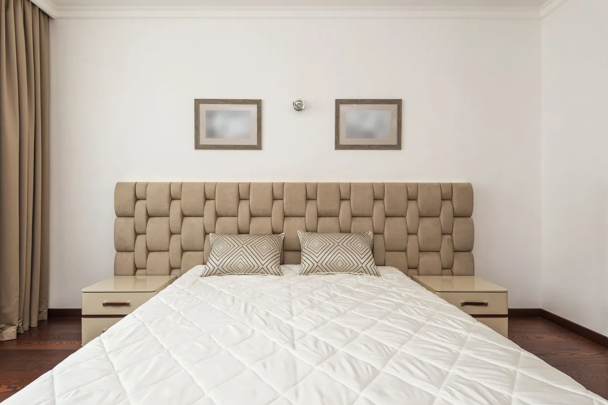 A bed with a white quilted cover, two patterned cushions and an upholstered headboard between two bedside tables