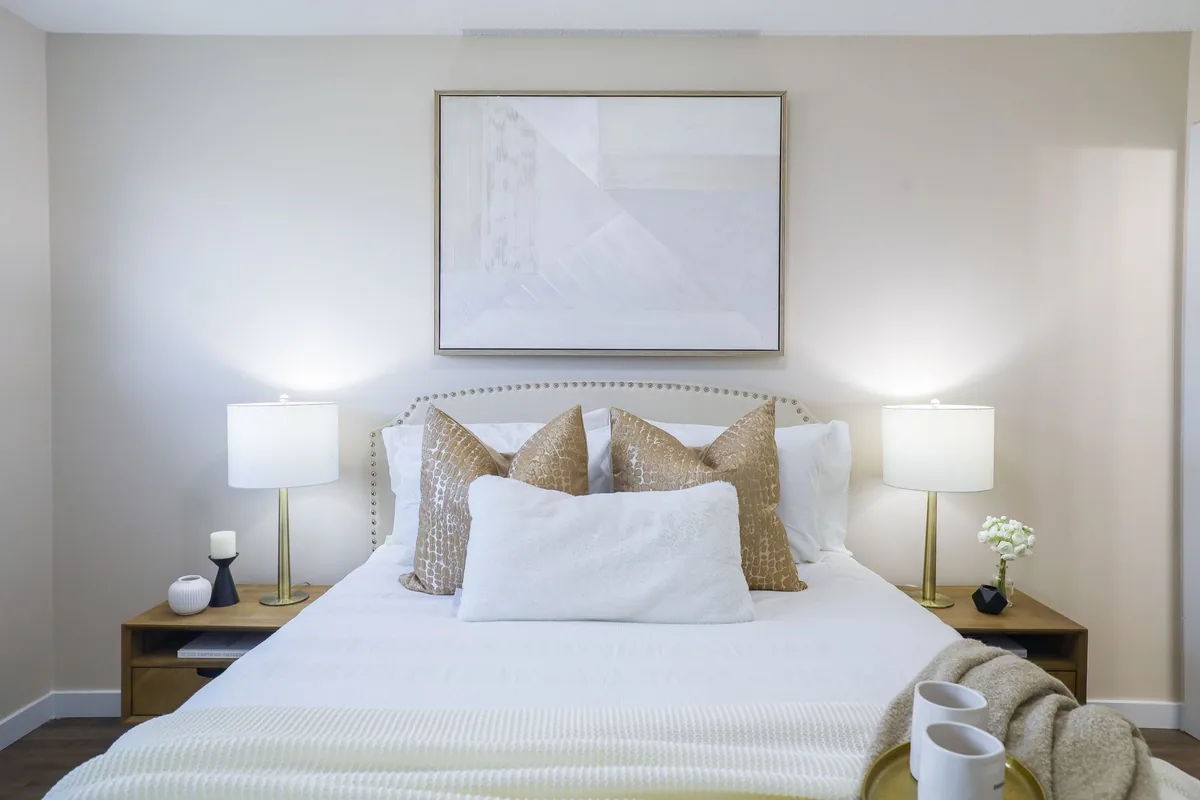A made bed centered under a large framed abstract painting, with a table lamp and wooden nightstand on each side and cushions against the headboard