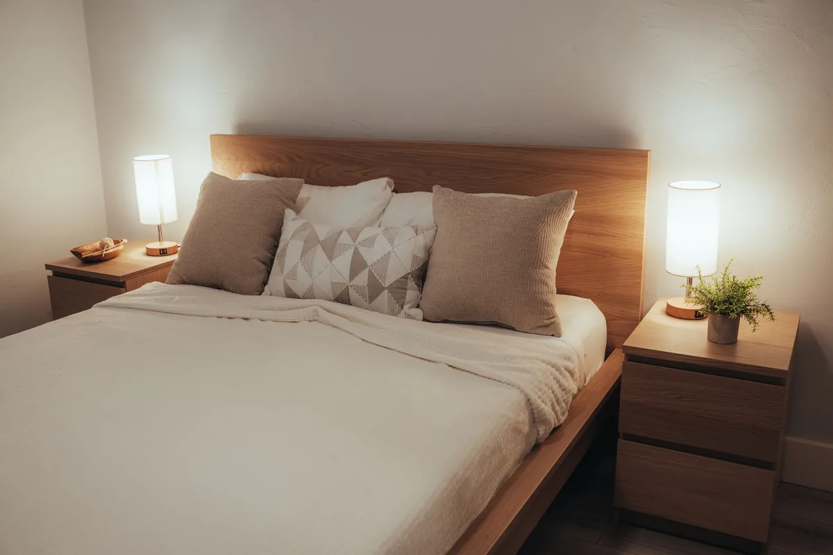 A bed with a pale wooden headboard, two large beige square cushions and a patterned cushion in front of white pillows, between two small table lamps on nightstands