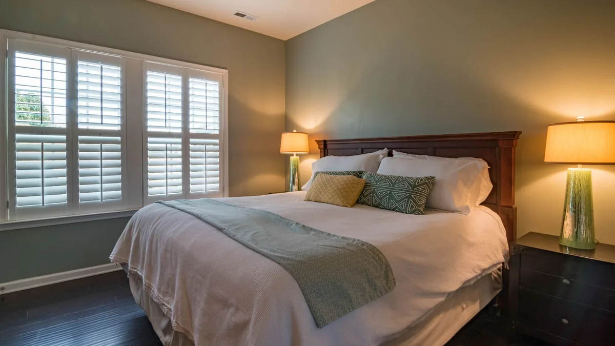 A bedroom with a bed dressed in white linens, a teal throw across the foot, two pillows, and lit table lamps on dark nightstands beside a window with white shutters