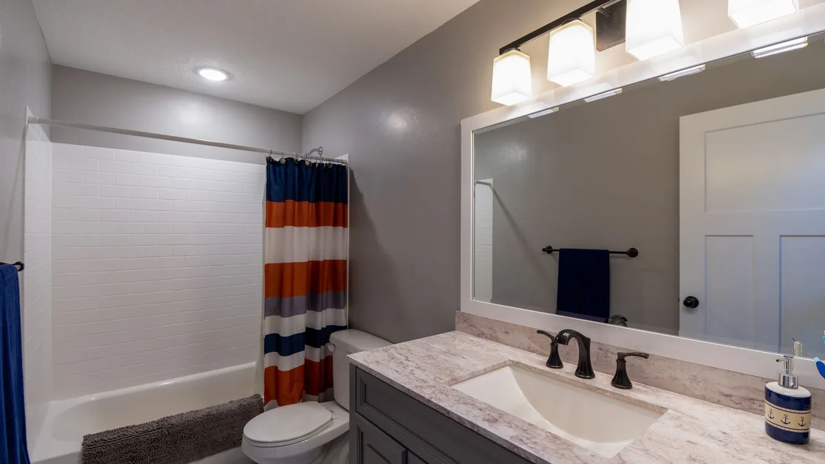 A bathroom with gray painted walls, white subway tile around the tub, a framed mirror and a row of vanity lights above a marble-look countertop