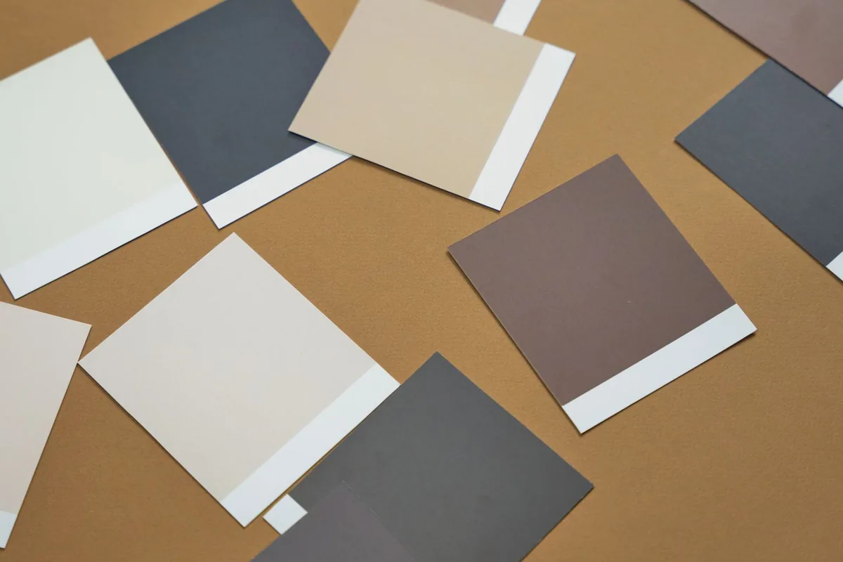 Paint color swatch cards in white, cream, beige, gray and brown scattered on a tan surface
