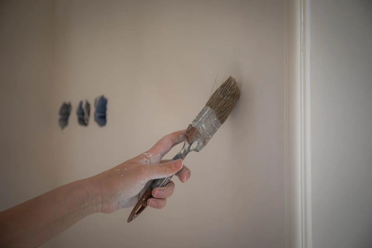 A hand brushing a large test patch onto a beige wall beside three small blue paint swatches