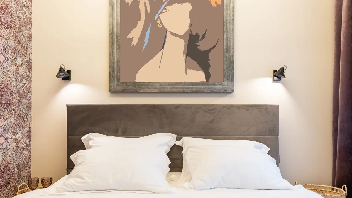 A made bed with white pillows against a gray upholstered headboard, a cream-painted wall, two black wall lamps and a framed print