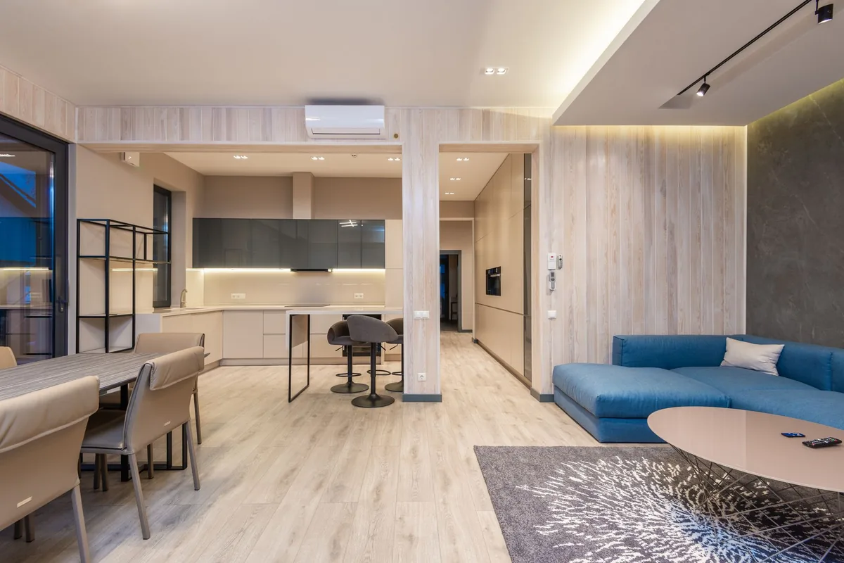 An open-plan room with a blue corner sofa, a gray rug, a dining table and a kitchen beyond a wood-paneled column