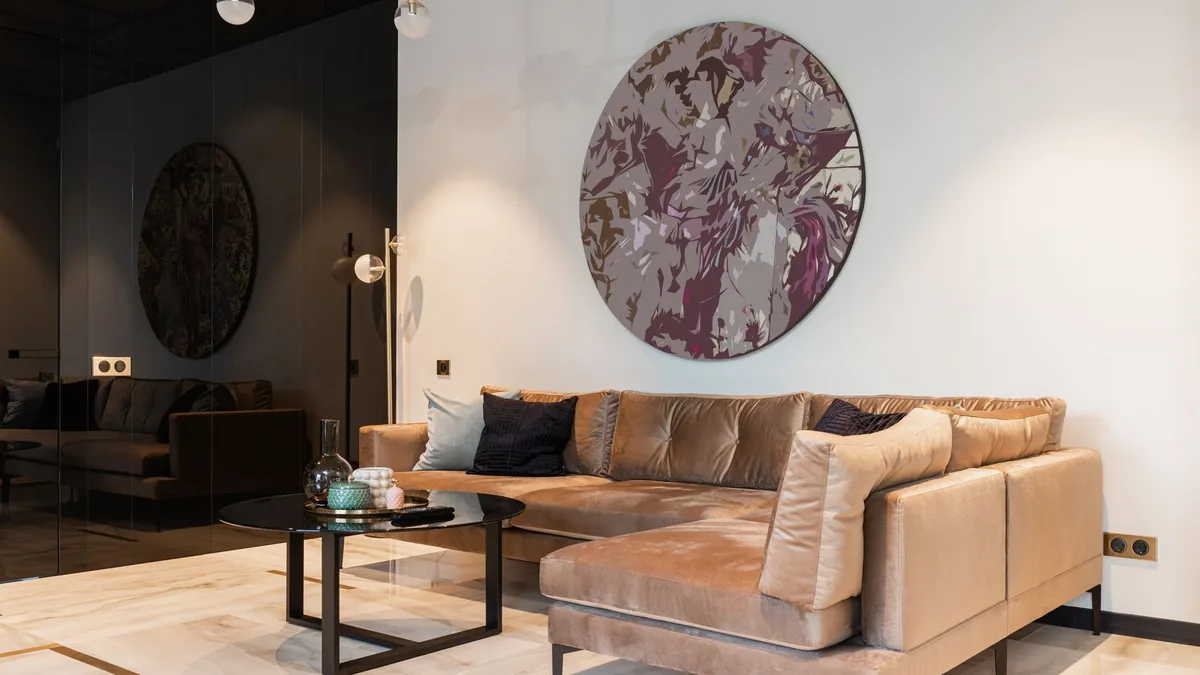 A living room with a tan velvet corner sofa against a white wall, a round wall artwork, a floor lamp and a glass-top coffee table on a pale marble floor