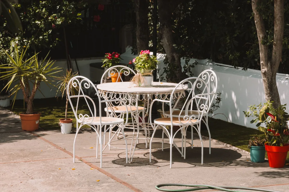A white metal patio table and chairs in dappled sun beside a garden wall, with potted plants around them