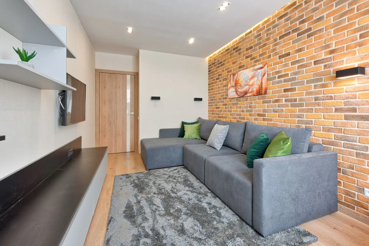 A grey chaise sectional along a brick wall in a narrow living room, with a grey rug and a wooden door at the far end