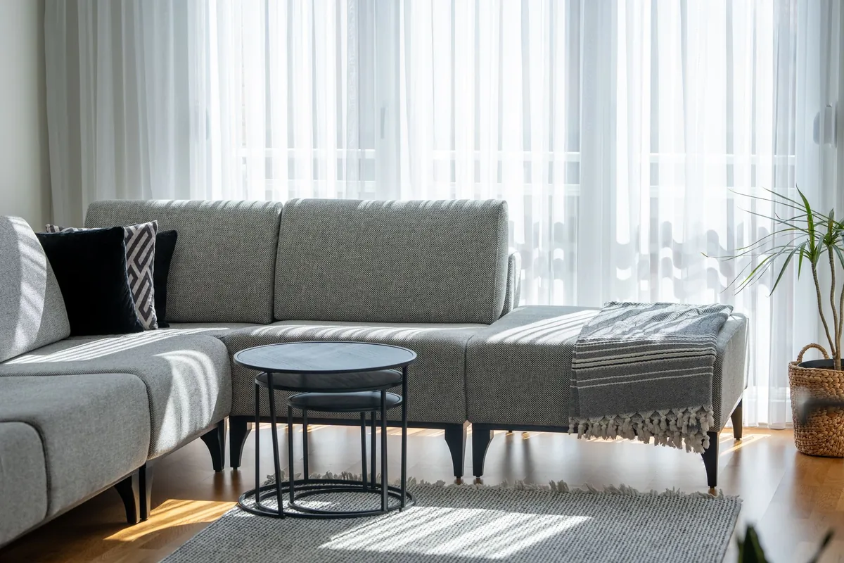 A grey corner sectional with black legs in front of sheer curtains, with a small round table and a patterned rug
