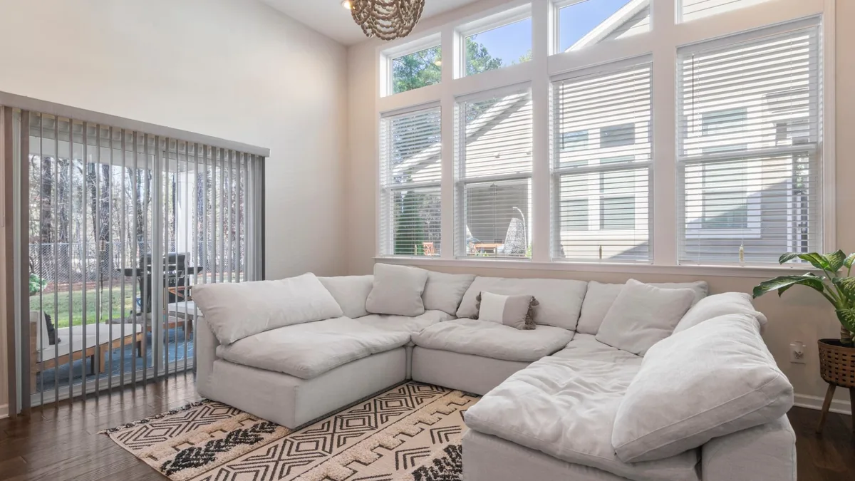 A light modular sectional in the corner of a living room under tall windows, with a patterned rug and a potted plant