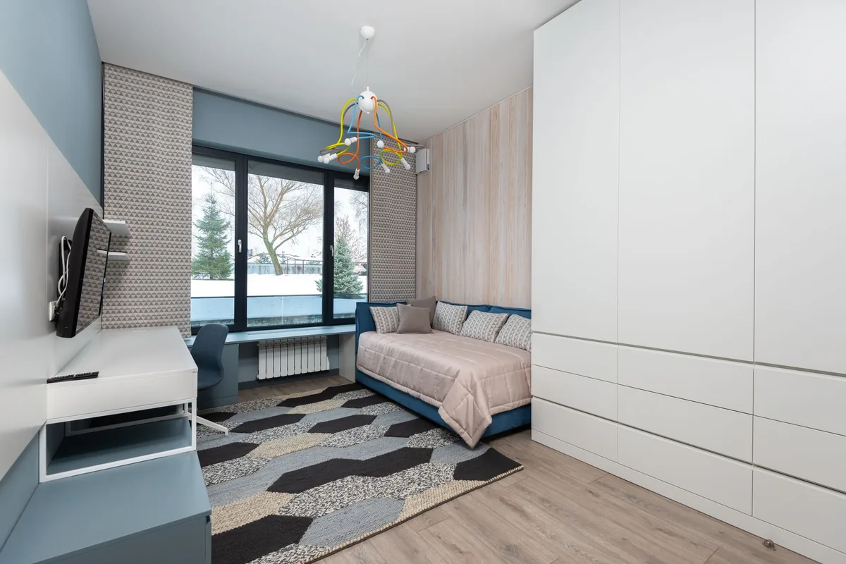 A bedroom with a blue upholstered daybed made up with a pale quilt and cushions, a desk under a wide window and a patterned rug