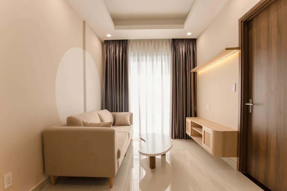 A narrow living room with a beige two-seat sofa, a small oval coffee table and a wall-mounted wooden media shelf