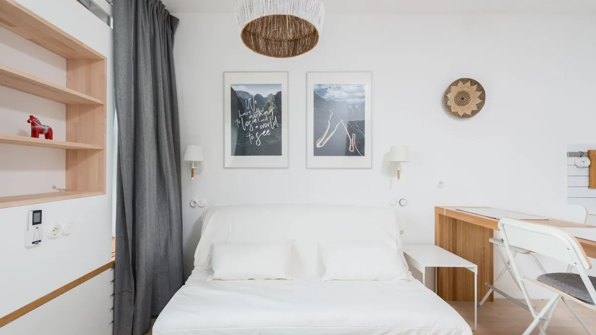An opened sofa bed made up with white bedding and two pillows in a bright studio, with wooden shelves, a desk and two framed prints