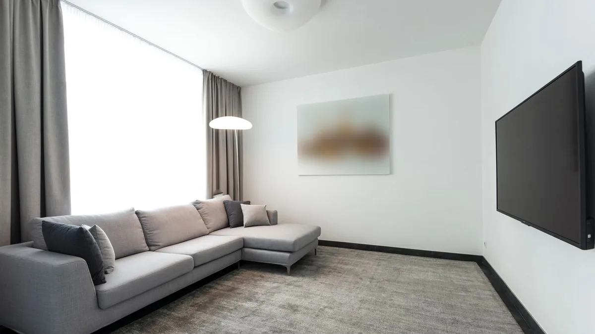 A bright modern living room with an L-shaped grey sofa on a large grey low-pile rug and a wall-mounted TV