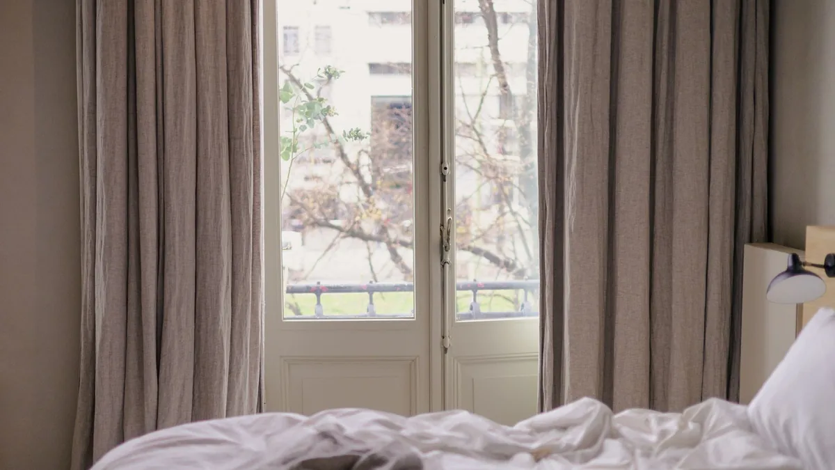Grey-beige curtains framing a glass balcony door beside an unmade bed with white sheets
