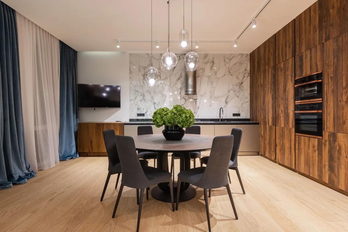 A round dining table on a pedestal base with six dark upholstered chairs in an open kitchen