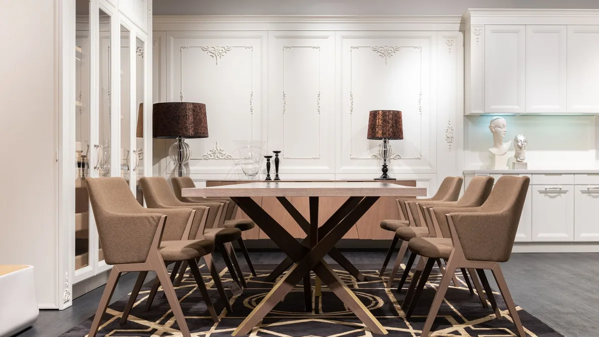 A long wooden dining table on a crossed base with eight upholstered armchairs, on a patterned rug in a white-paneled room