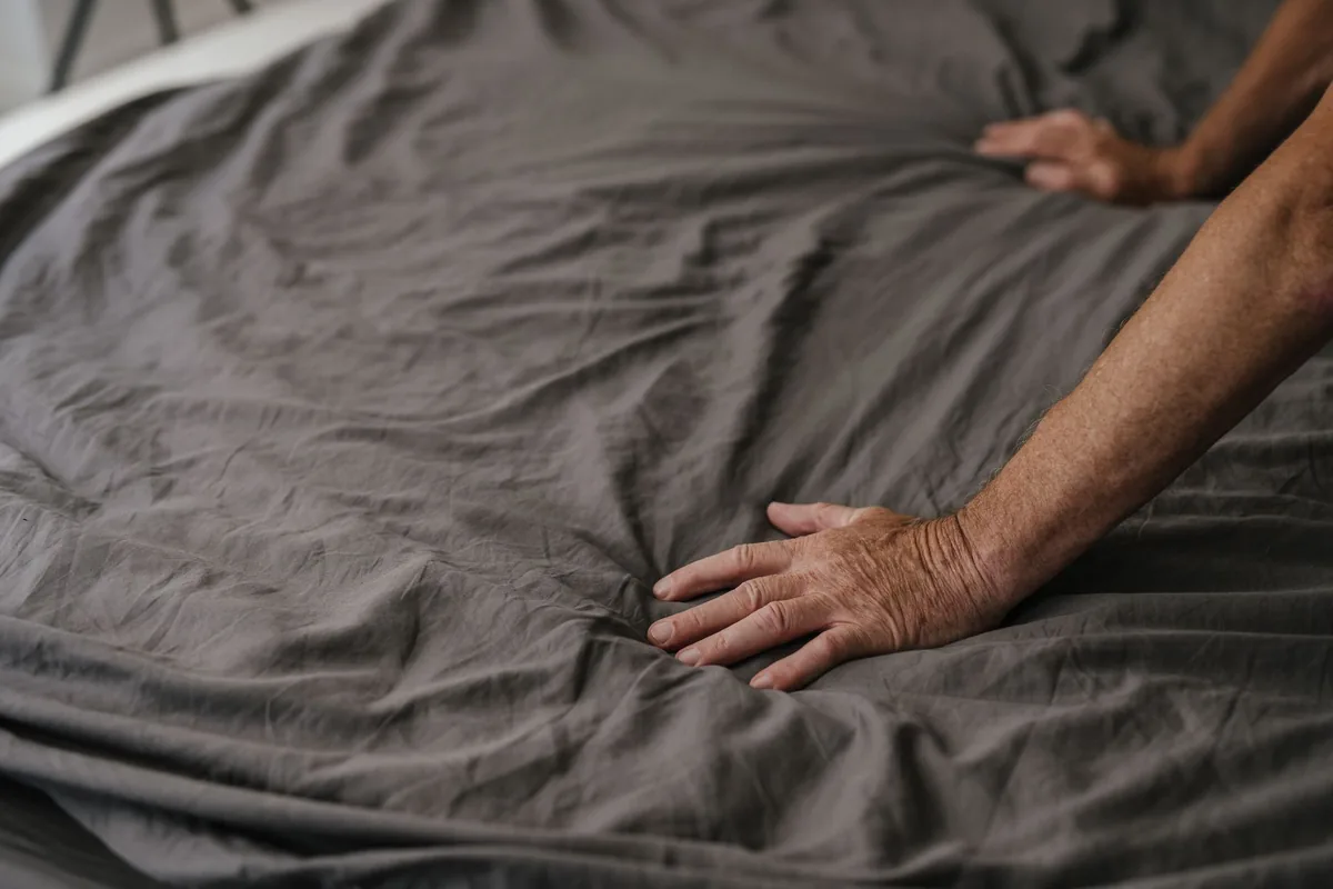 Hands smoothing a wrinkled grey cover on a bed