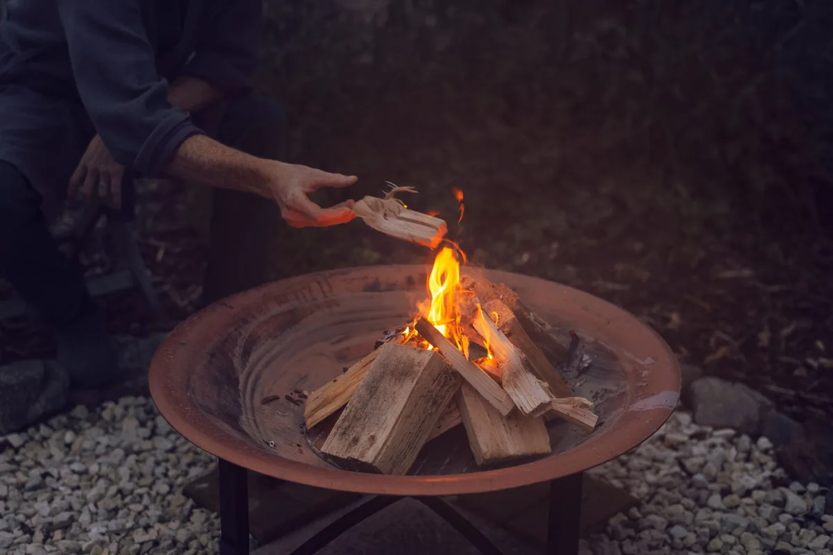 A person adding split firewood to a small fire in a round steel fire bowl standing on gravel