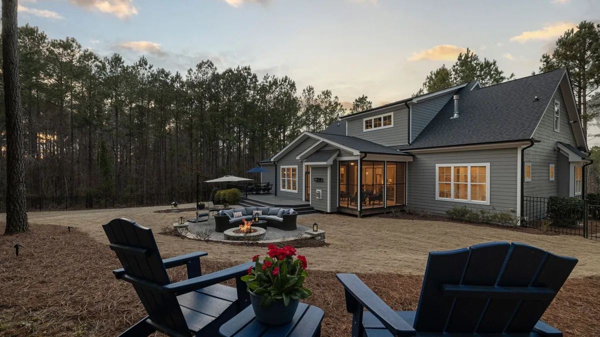A backyard at dusk with a round stone fire pit on a paved patio, a curved outdoor sofa beside it and two Adirondack chairs in the foreground