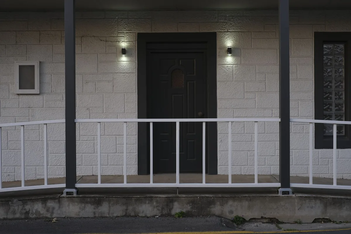 A dark front door on a covered porch with a small lit wall light on each side and a white railing in front