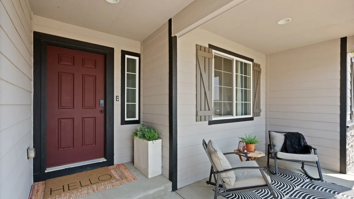 A covered front porch with a red front door, a door mat, a tall white planter beside the door and two cushioned rocking chairs on a patterned rug