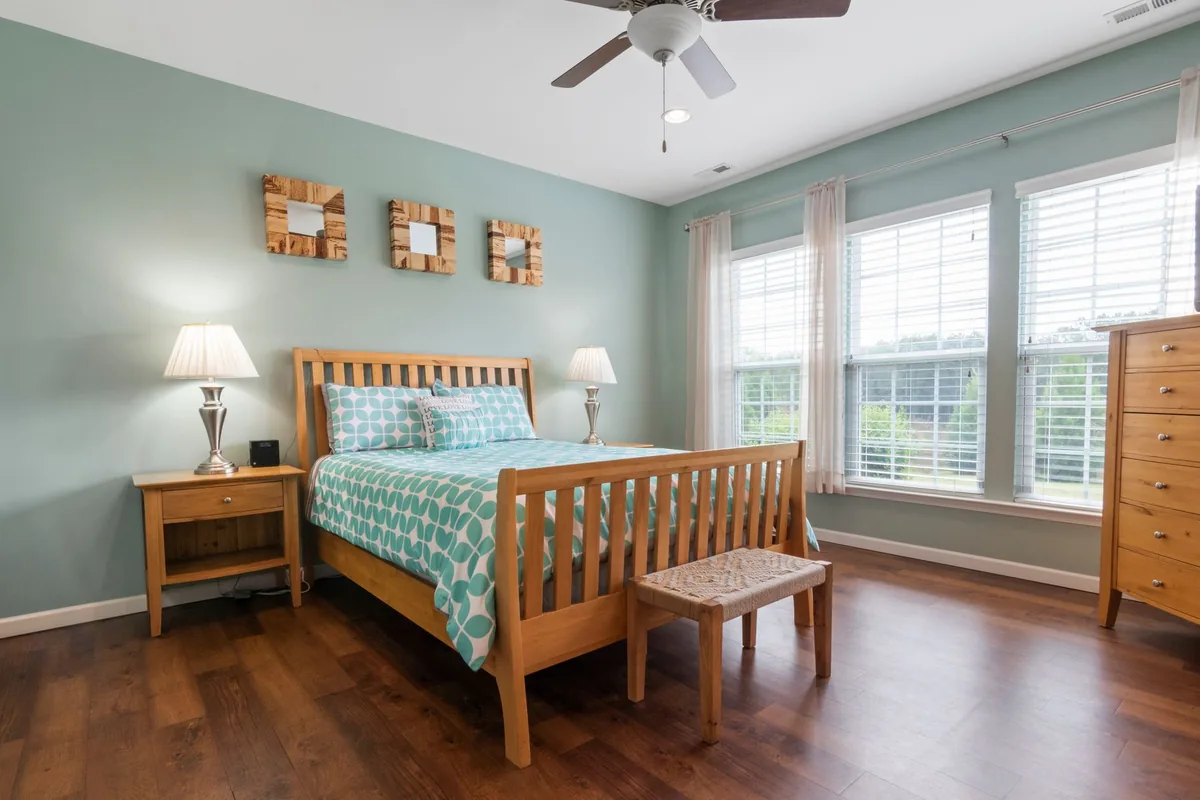 A bedroom with a slatted wooden bed between two wooden nightstands with lamps, and a tall chest of drawers by the windows