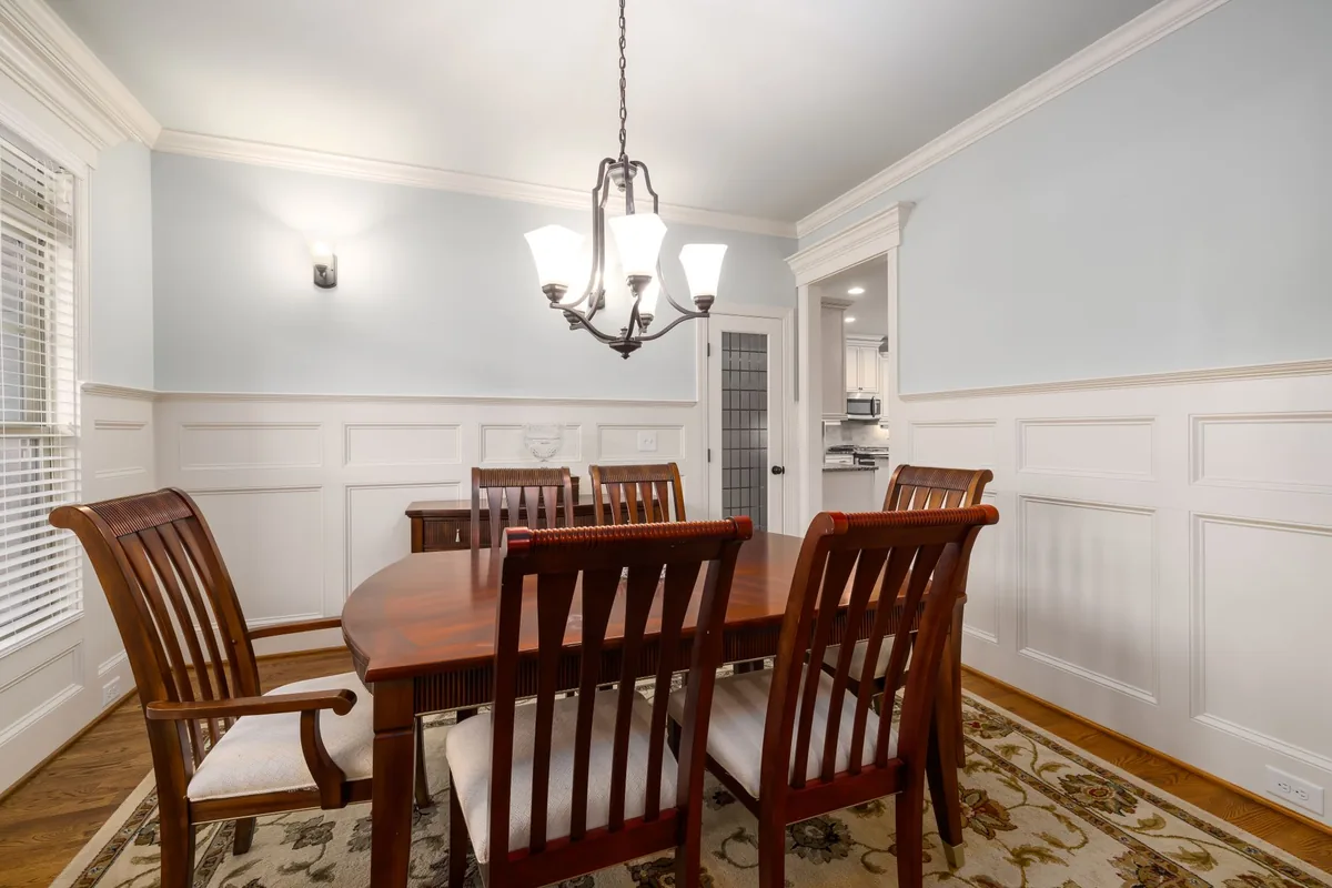 A dining room with a wooden table and slat-back wooden chairs on a patterned rug under a pendant light