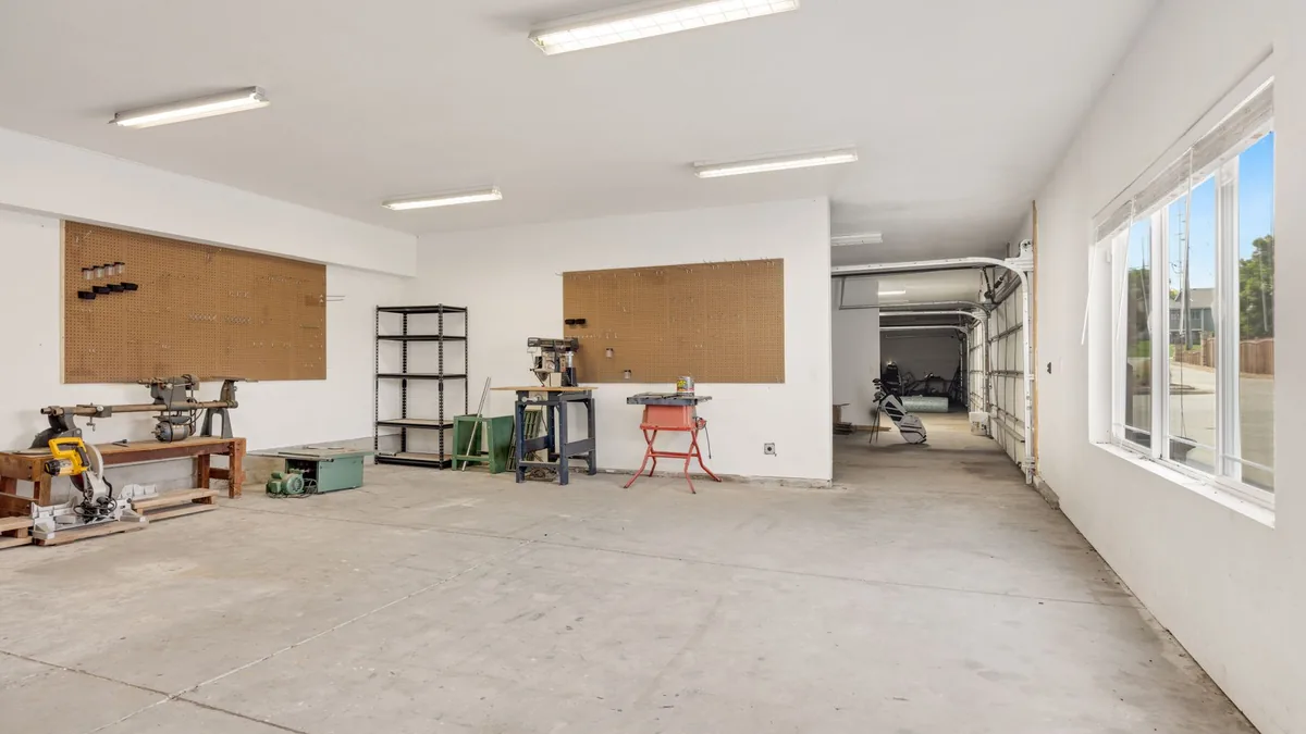 A bright, mostly empty garage with pegboard on two walls, an open metal shelving unit, a workbench and an open garage door at the far end