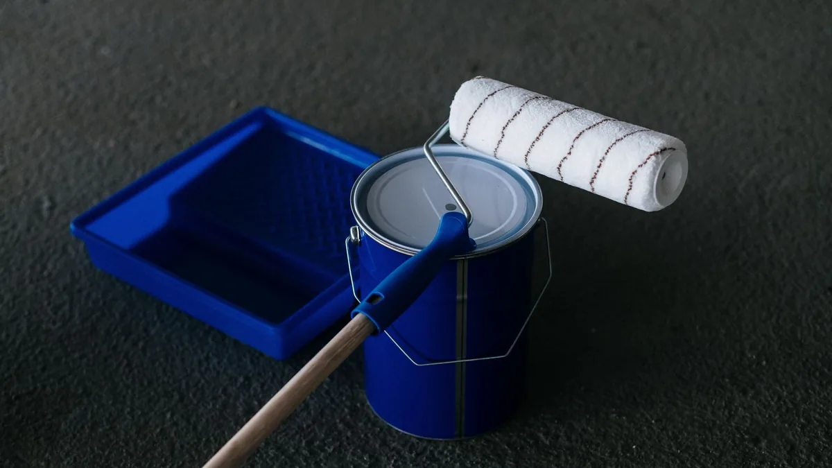 A paint roller resting on a closed paint can next to an empty roller tray on a dark concrete floor