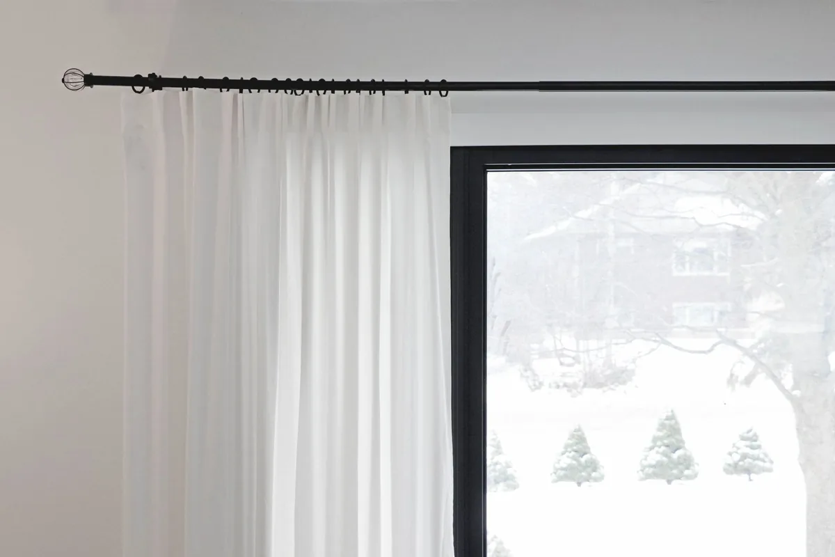 A black curtain rod with rings and a finial running well past the edge of a window, with a white pleated curtain hanging beside the glass
