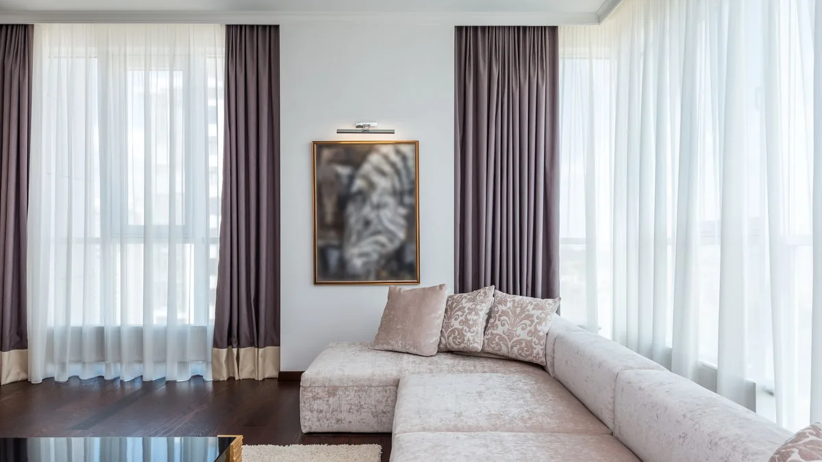 A bright living room with floor-length sheer curtains and mauve drapes hung high on tall windows, a pale sectional sofa and a framed picture between the windows