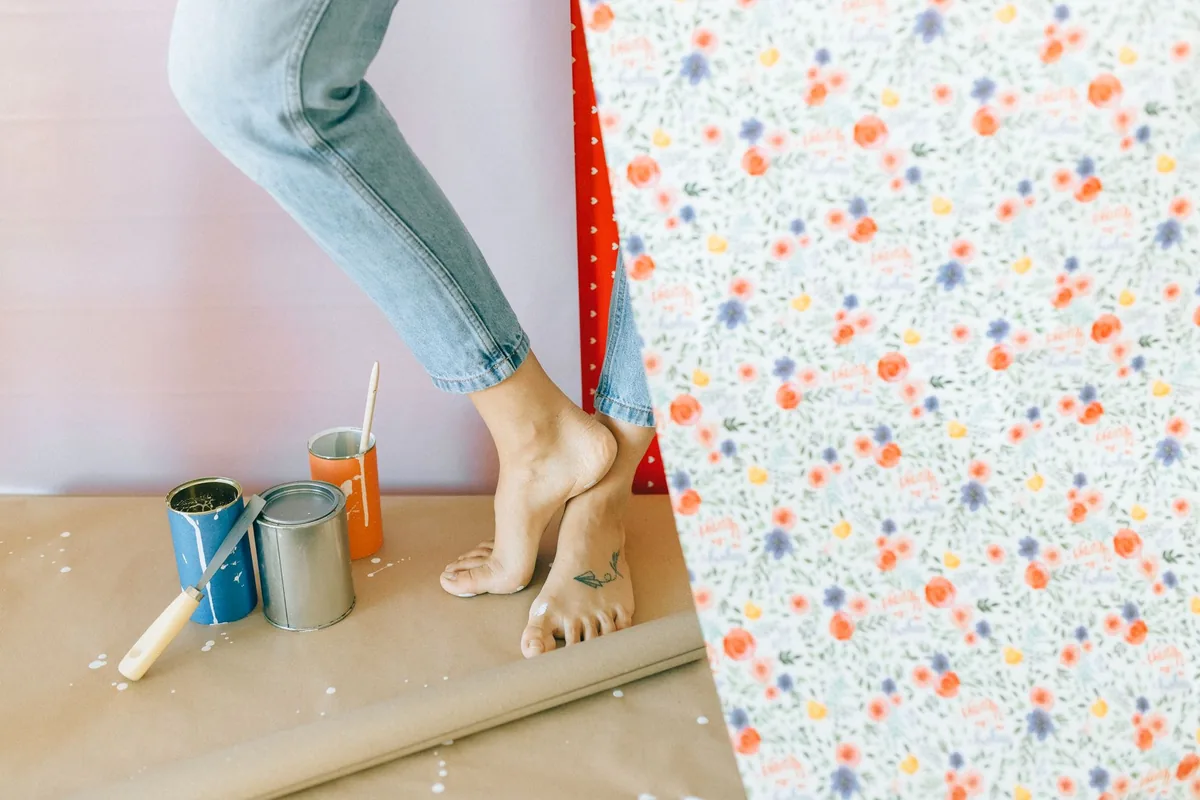 A person standing barefoot on brown paper next to open paint cans, holding up a roll of floral wallpaper