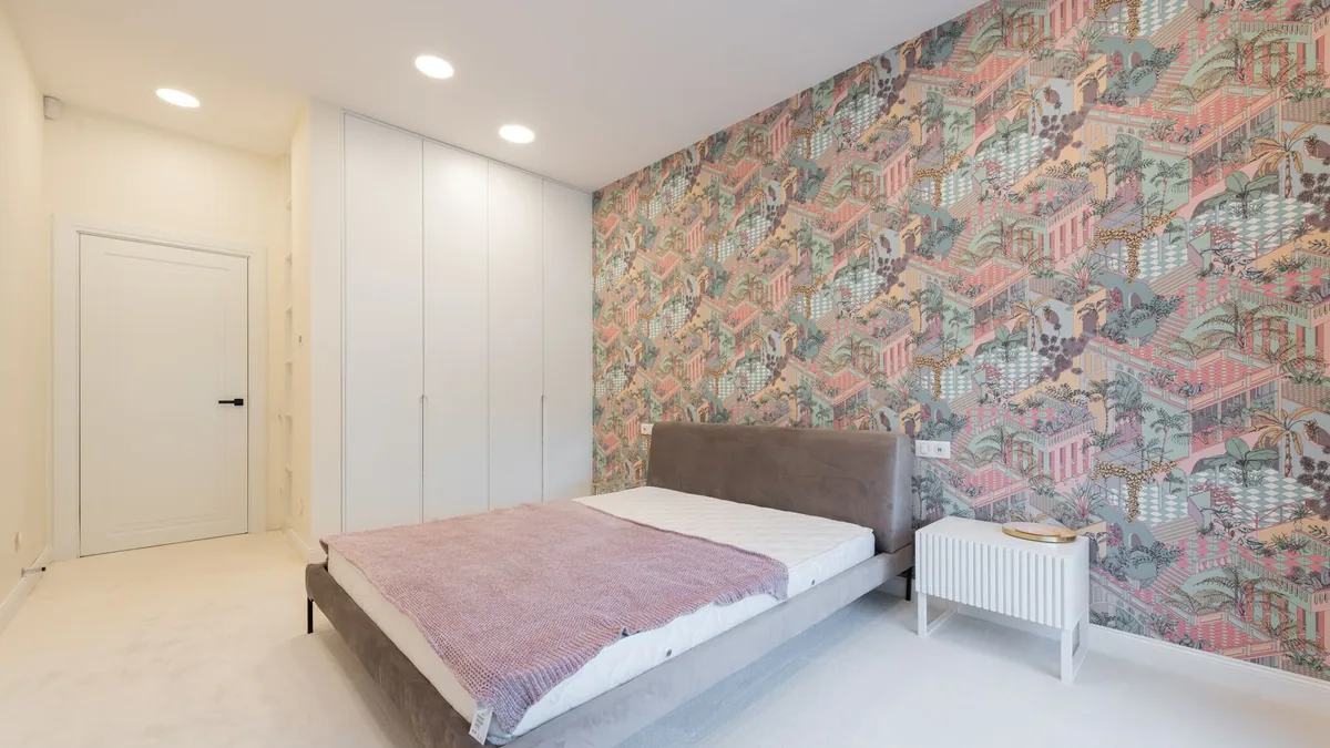 A bedroom with one wall covered in pink, green and yellow patterned wallpaper behind a gray upholstered bed and a white nightstand