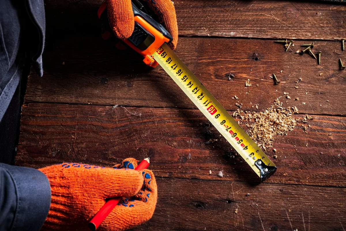 Gloved hands holding a pencil and a tape measure stretched across a wooden board