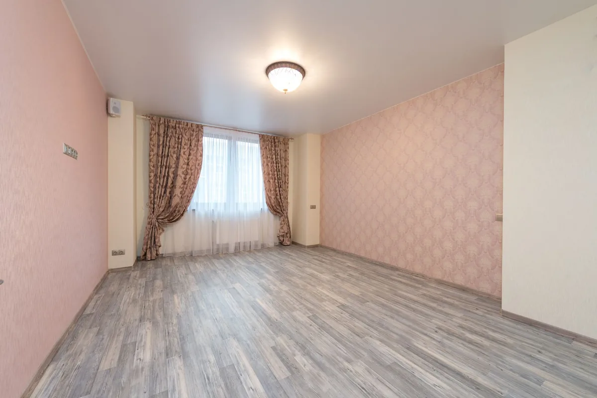 An empty room with pink patterned wallpaper, long curtains and a gray wood-look plank floor finished with baseboards