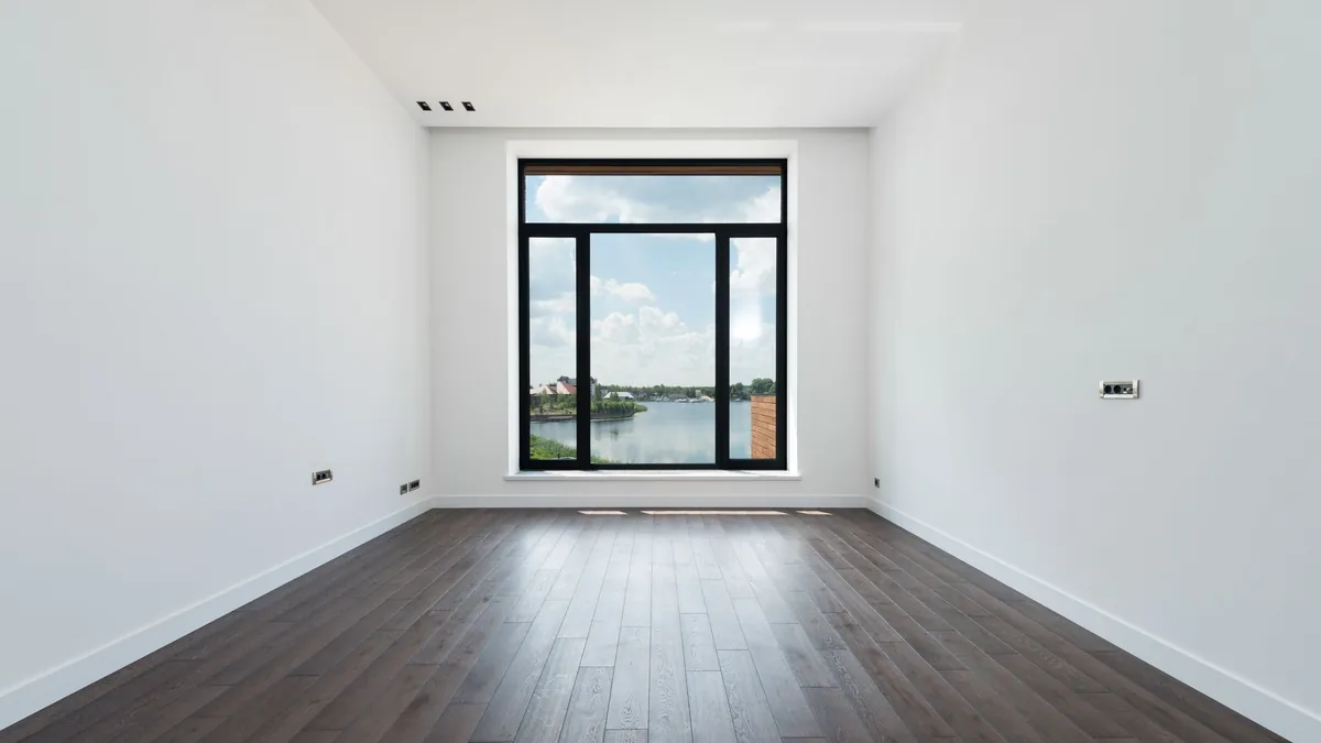 An empty room with white walls, a large window and a dark wood-look plank floor running toward the window