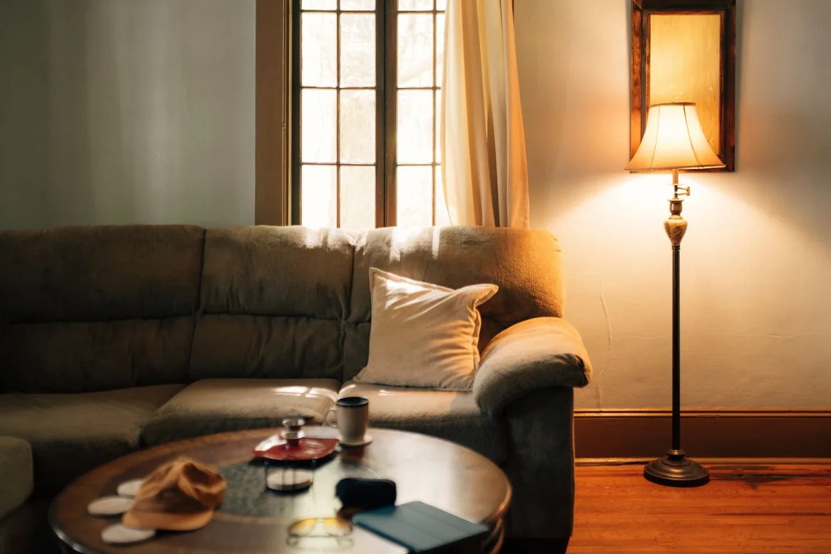 A floor lamp with a fabric shade glowing beside a sofa and a round coffee table near a window