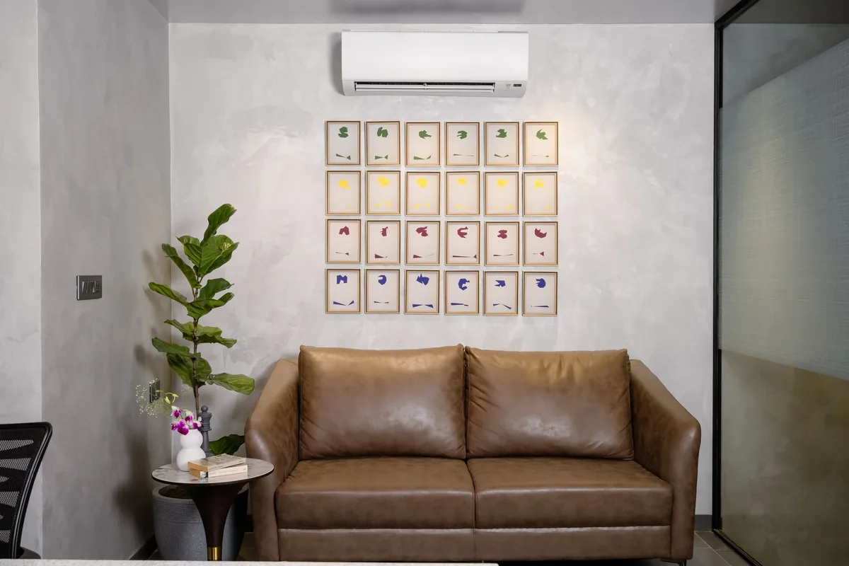 A grid of small framed prints centered on the wall above a brown leather loveseat, with a side table and a potted plant