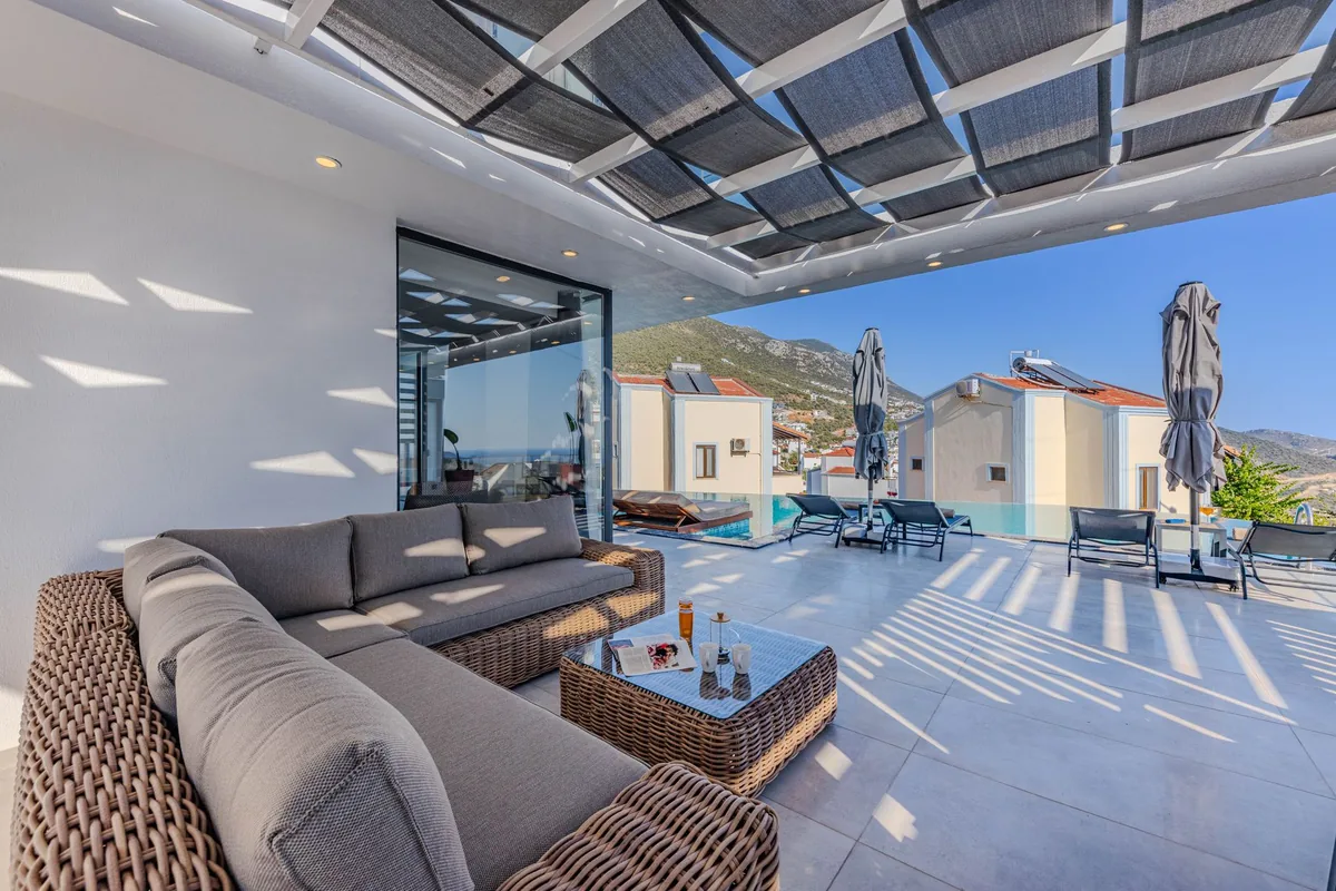An L-shaped wicker outdoor sofa with grey cushions and a glass-top wicker coffee table on a large covered terrace beside a pool