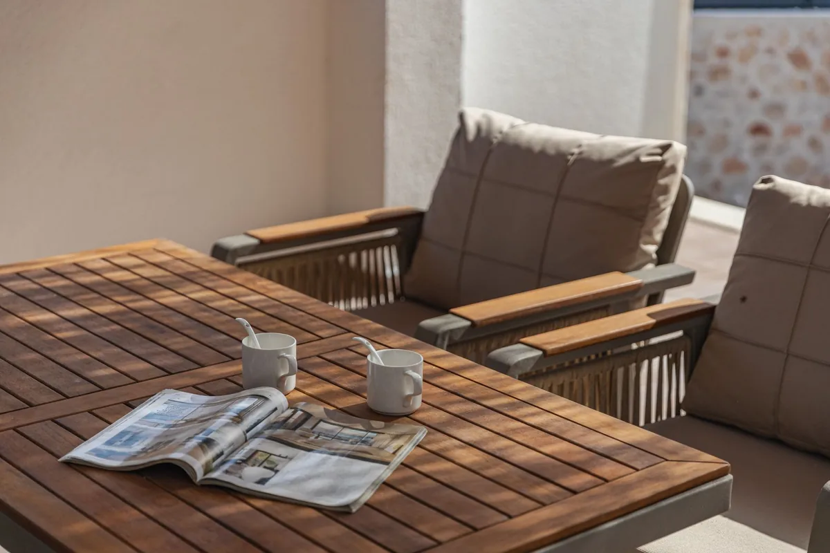 Two cushioned armchairs with wooden armrests beside a slatted wooden table with a magazine and two cups on a sunny patio