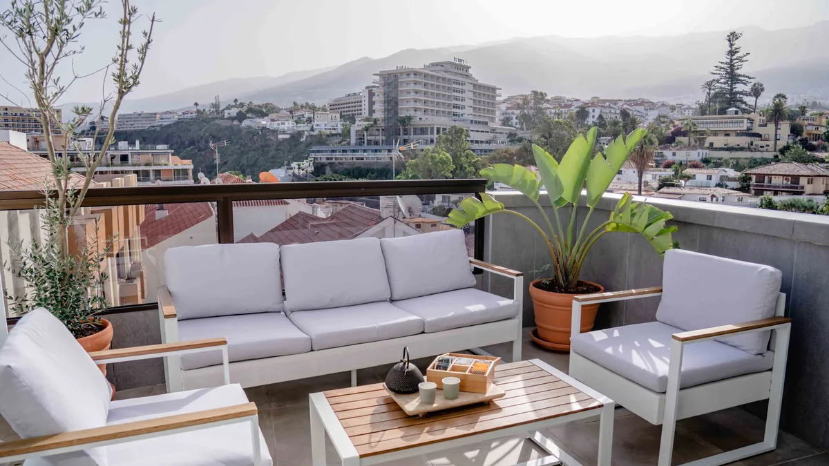 A balcony lounge area with a three-seat sofa, two armchairs with grey cushions and a wood-top coffee table, with hills and rooftops beyond the railing